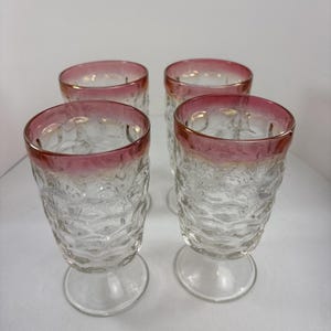 May include: A set of four clear glass goblets featuring a textured honeycomb pattern on the bowl and a distinct pink rim. Each glass stands on a circular base, showcasing a clear stem and a textured body that catches the light with its unique design.