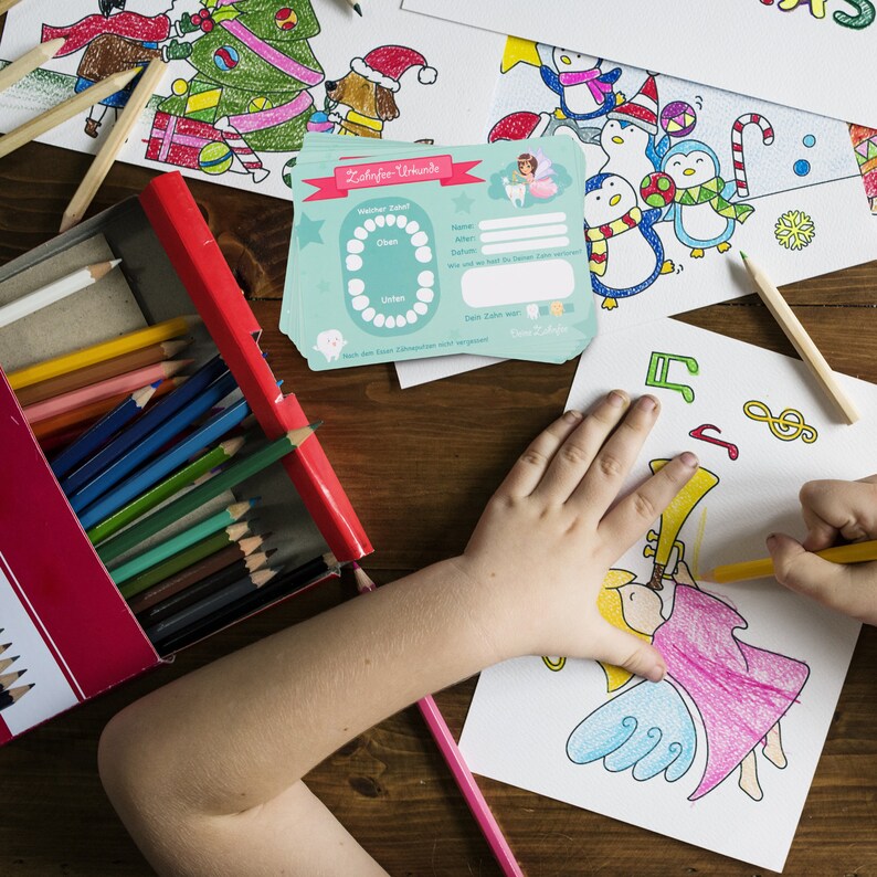 May include: A turquoise blue card with the title "Z&auml;hltafel - Vorlage" and a chart for tracking lost teeth. The card has spaces for the child's name, age, date, and the name of the lost tooth. The card is on a wooden table with a box of colored pencils and a drawing of a child coloring a picture of an angel with a trumpet.