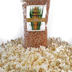 May include: A bag of yellow popcorn kernels with a label that reads "Meyer's Homegrown - Premium. Yellow Popcorn. All Natural - Whole Grain - Non GMO - Gluten-Free. Net Wt 2 lbs (907 g)". A pile of popped popcorn is in the foreground.