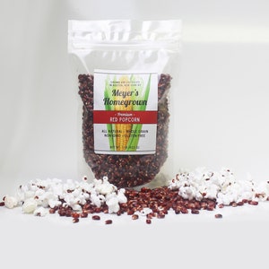May include: A clear plastic bag filled with red popcorn kernels. The bag has a label that reads "Meyer's Homegrown - Premium - Red Popcorn - All Natural - Whole Grain - Non-GMO - Gluten-Free - Net Wt 1 lb (453 g)". There are also popped red popcorn kernels scattered around the bag.