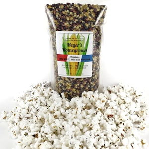 May include: A bag of red, white, and blue popcorn kernels. The bag is labeled "Meyer's Homegrown Premium Red, White, and Blue Popcorn". The label also states that the popcorn is all natural, whole grain, non-GMO, and gluten-free. The bag is sitting on a pile of popped popcorn.