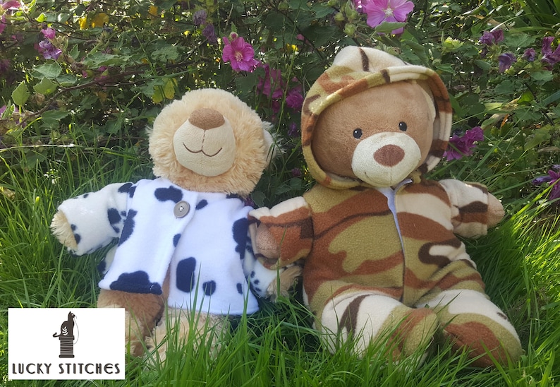 May include: Two plush teddy bears sit in the grass. One bear wears a white jacket with black spots. The other bear is dressed in a camouflage-patterned outfit with a hood. The background features green grass and purple flowers.