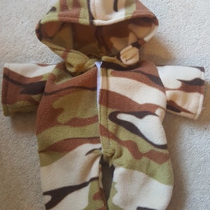 May include: A hooded camouflage-patterned onesie with a full-length zipper. The onesie features a mix of brown, green, and cream-colored fabric. The hood has small ear-like details. The garment is designed for a baby or toddler.