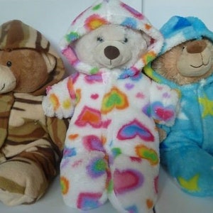 May include: Three plush teddy bears wearing hooded outfits. One bear is in a camouflage pattern, another in a white onesie with colorful hearts, and the third in a blue outfit with yellow stars. The bears are soft and cuddly.