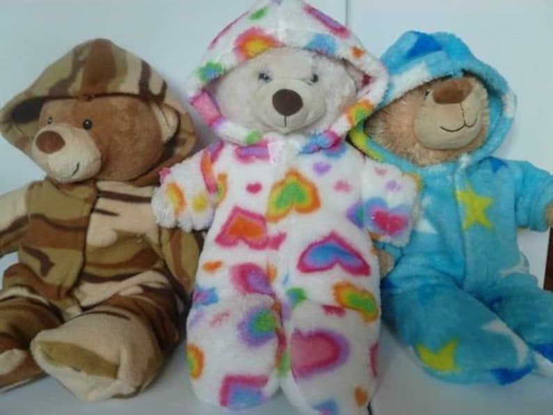 May include: Three plush teddy bears wearing hooded outfits. One bear is in a camouflage pattern, another in a white onesie with colorful hearts, and the third in a blue outfit with yellow stars. The bears are soft and cuddly.