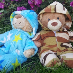 May include: Two teddy bears in a grassy area with purple flowers. One bear wears a blue onesie with star patterns. The other bear is dressed in a camouflage-patterned outfit. Both bears have hoods.