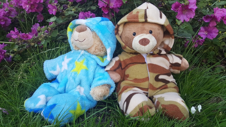 May include: Two teddy bears in a grassy area with purple flowers. One bear wears a blue onesie with star patterns. The other bear is dressed in a camouflage-patterned outfit. Both bears have hoods.