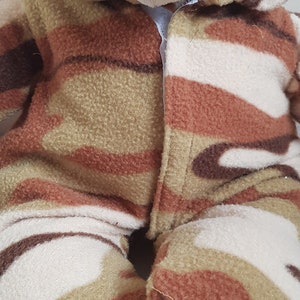 May include: A plush teddy bear wearing a camouflage-patterned outfit in shades of brown, beige, and olive green. The bear has a light brown head, a white muzzle, and black eyes. The outfit covers the bear's body and includes a hood.