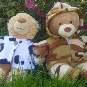May include: Two plush teddy bears sit in the grass. One bear wears a white coat with navy blue spots. The other bear is dressed in a camouflage-patterned outfit with a hood. The bears are surrounded by green grass and purple flowers.
