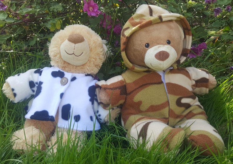 May include: Two plush teddy bears sit in the grass. One bear wears a white coat with navy blue spots. The other bear is dressed in a camouflage-patterned outfit with a hood. The bears are surrounded by green grass and purple flowers.