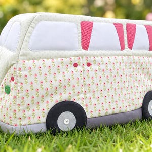 May include: A quilted fabric cover shaped like a camper van, featuring a light green background with a repeating pink floral pattern. The design includes white windows, pink polka-dot curtains, dark gray wheels with white buttons, and green accents.