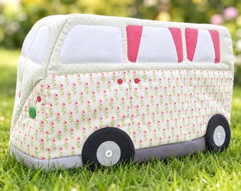 May include: A quilted fabric cover shaped like a camper van, featuring a light green background with a repeating pink floral pattern. The design includes white windows, pink polka-dot curtains, dark gray wheels with white buttons, and green accents.