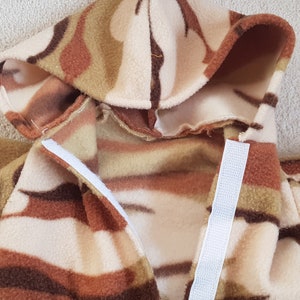 May include: A hooded jacket made of soft, patterned fleece. The jacket features a camouflage design in shades of brown, tan, and cream. White Velcro strips secure the front closure. The jacket has a hood and sleeves.