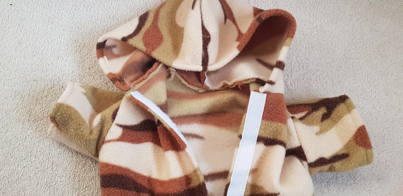 May include: A hooded jacket made of soft, patterned fleece. The jacket features a camouflage design in shades of brown, tan, and cream. White Velcro strips secure the front closure. The jacket has a hood and sleeves.