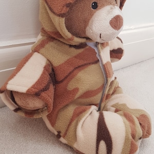 May include: A brown teddy bear wearing a camouflage-patterned hooded jumpsuit. The jumpsuit features a zipper closure and covers the bear's body, arms, and legs. The bear is sitting on a light-colored carpet.