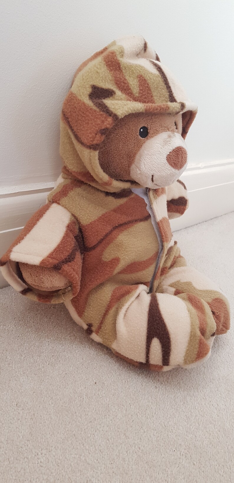May include: A brown teddy bear wearing a camouflage-patterned hooded jumpsuit. The jumpsuit features a zipper closure and covers the bear's body, arms, and legs. The bear is sitting on a light-colored carpet.