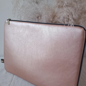 Peut inclure: Une housse d'ordinateur portable rose blush avec un aspect texturé semblable au cuir. La housse a une fermeture éclair noire et une petite étiquette de couleur foncée sur le côté. Le fond présente des herbes décoratives duveteuses et de couleur claire.