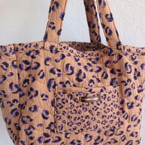 Könnte beinhalten: Eine gesteppte Tragetasche in Beige mit einem lila-schwarzen Leopardenmuster. Die Tasche verfügt über zwei Schulterriemen und eine Vordertasche mit einem braunen rechteckigen Etikett, auf dem der Text FREAKS & BAGS in Weiß gedruckt ist.