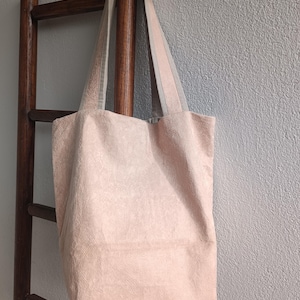 Pode incluir: Uma bolsa tote rosa pálido feita de um tecido texturizado semelhante a uma toalha. A bolsa possui duas alças de ombro longas e combinando, presas à borda superior. O material tem uma textura de superfície visível, ligeiramente elevada e em laços.
