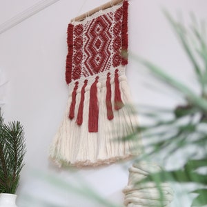 Peut inclure: Une tapisserie murale tissée blanche et rouge avec des glands. La tapisserie présente un motif géométrique et est faite de fibres naturelles.