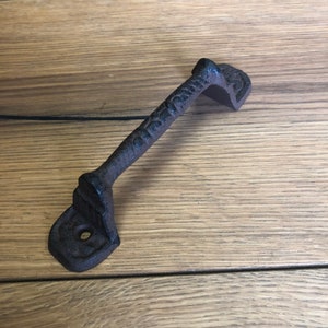 May include: A black cast iron door pull with a decorative design. The pull is mounted on a wooden surface.