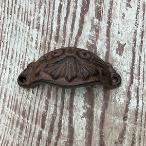 May include: A brown cast iron drawer pull with a decorative floral design. The pull is mounted on a whitewashed wooden surface.