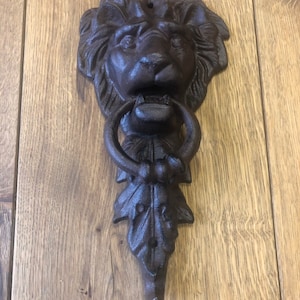 May include: A cast iron door knocker in the shape of a lion's head. The knocker is dark brown with a ring for knocking and a decorative leaf design. The lion's face has detailed features.