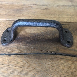 May include: Black cast iron door handle with a rounded rectangular shape. The handle has two holes on each side for mounting.