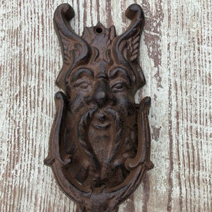 May include: A dark brown cast iron door knocker featuring a detailed relief of a bearded face with prominent horns or leafy protrusions at the top. The heavy metal object has a hinged U-shaped ring for knocking and a mounting hole at the top.