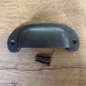 May include: A black metal drawer pull with a curved shape and two screw holes. The pull is on a wooden surface with two black screws.