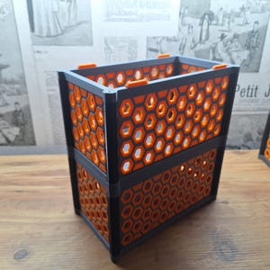 May include: A two-tiered, rectangular container featuring a honeycomb design. The container is black with orange honeycomb inserts. The design is geometric and modern. The background includes a wooden surface and a vintage newspaper.