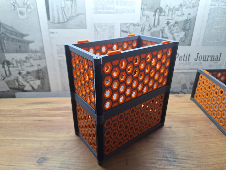 May include: A two-tiered, rectangular container featuring a honeycomb design. The container is black with orange honeycomb inserts. The design is geometric and modern. The background includes a wooden surface and a vintage newspaper.