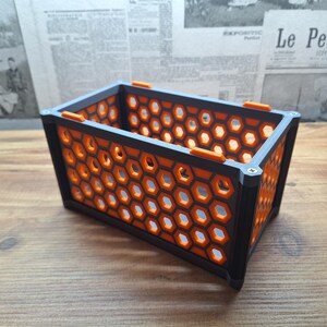 以下が含まれることがあります： ハニカム模様の長方形収納容器。容器は黒色で、オレンジ色のアクセントと灰色のフレームが付いています。ハニカムデザインは光を通します。容器は木製の表面にあります。