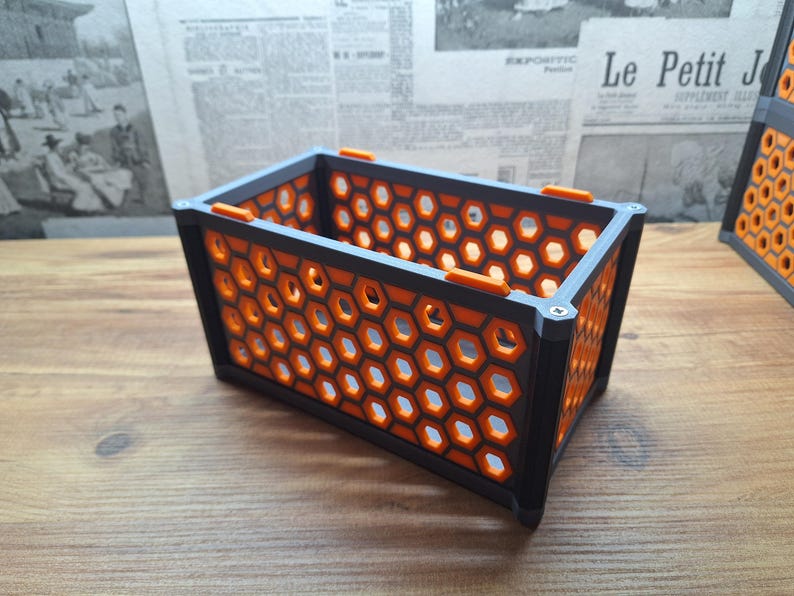 May include: A rectangular storage container with a honeycomb pattern. The container is black with orange accents and a grey frame. The honeycomb design allows light to pass through. The container is on a wooden surface.