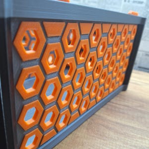 May include: A rectangular, black and orange decorative box with a honeycomb pattern. The box features a series of orange hexagonal cutouts set against a dark grey background. The top and sides are black, creating a striking contrast. The box is on a wooden surface.