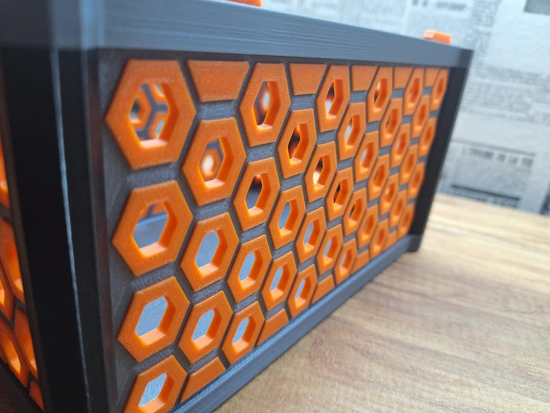 May include: A rectangular, black and orange decorative box with a honeycomb pattern. The box features a series of orange hexagonal cutouts set against a dark grey background. The top and sides are black, creating a striking contrast. The box is on a wooden surface.