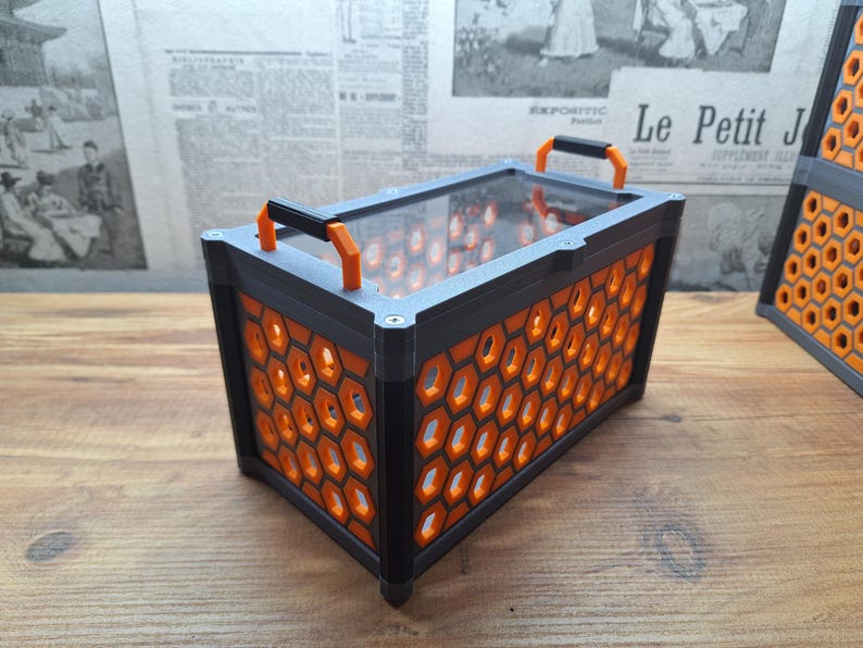 May include: Rectangular black and orange container with a clear top and orange honeycomb pattern sides. The container has two orange handles and is sitting on a wooden surface. The container is approximately 25 cm long.