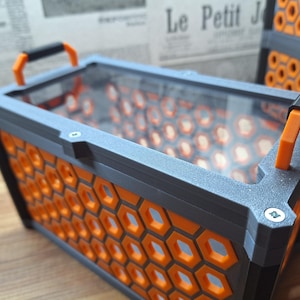 May include: A rectangular container with a grey frame and orange honeycomb pattern sides. It has a clear top and orange handles. The container is on a wooden surface.