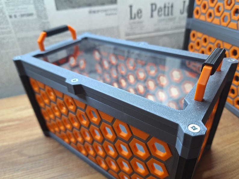 May include: A rectangular container with a grey frame and orange honeycomb pattern sides. It has a clear top and orange handles. The container is on a wooden surface.