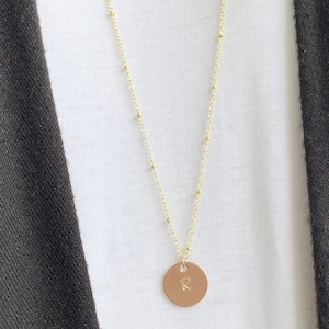 May include: A gold-colored necklace with a delicate chain and small beaded accents. The necklace features a round, tan pendant engraved with the letter "R". The necklace is worn over a white shirt and a dark gray cardigan.