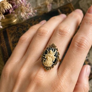 K&ouml;nnte beinhalten: Ein goldfarbener Ring mit einem ovalen Cameo, der eine cremefarbene Engelsfigur vor schwarzem Hintergrund zeigt. Der Ring hat einen verzierten Rahmen im Filigranstil. Der Ring befindet sich an einer Hand.