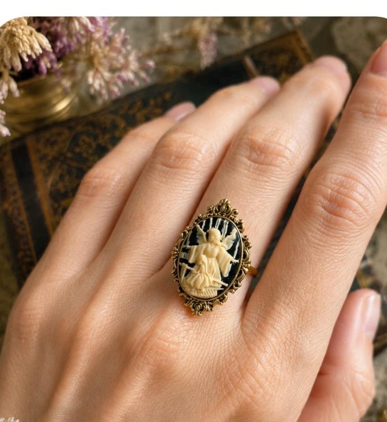 K&ouml;nnte beinhalten: Ein goldfarbener Ring mit einem ovalen Cameo, der eine cremefarbene Engelsfigur vor schwarzem Hintergrund zeigt. Der Ring hat einen verzierten Rahmen im Filigranstil. Der Ring befindet sich an einer Hand.