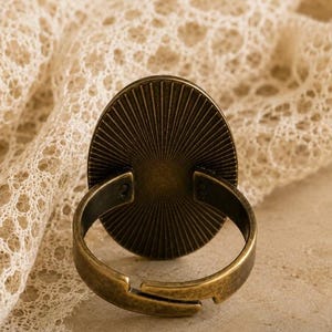 K&ouml;nnte beinhalten: Antiker, verstellbarer Ring in Bronzefarbe mit ovalem, sonnenstrahlenf&ouml;rmigem Gesicht. Der Ring ist vor einem Hintergrund aus cremefarbener Spitze platziert, was eine Vintage-&Auml;sthetik erzeugt. Design und Farbe des Rings deuten auf ein klassisches Schmuckst&uuml;ck hin.