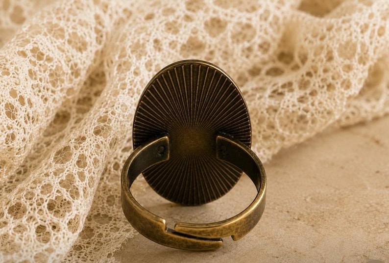K&ouml;nnte beinhalten: Antiker, verstellbarer Ring in Bronzefarbe mit ovalem, sonnenstrahlenf&ouml;rmigem Gesicht. Der Ring ist vor einem Hintergrund aus cremefarbener Spitze platziert, was eine Vintage-&Auml;sthetik erzeugt. Design und Farbe des Rings deuten auf ein klassisches Schmuckst&uuml;ck hin.
