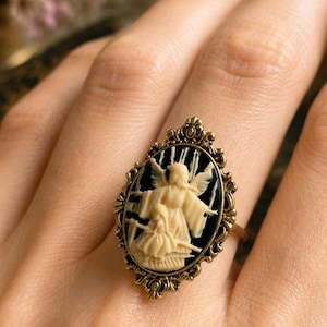K&ouml;nnte beinhalten: Antik anmutender Ring mit ovaler Kamee, die eine cremefarbene Engelsfigur vor schwarzem Hintergrund zeigt. Der Ring hat einen goldfarbenen Filigranrahmen und -band und pr&auml;sentiert eine Vintage-&Auml;sthetik. Der Ring wird am Finger getragen.