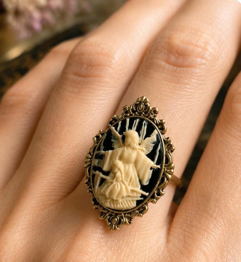 K&ouml;nnte beinhalten: Antik anmutender Ring mit ovaler Kamee, die eine cremefarbene Engelsfigur vor schwarzem Hintergrund zeigt. Der Ring hat einen goldfarbenen Filigranrahmen und -band und pr&auml;sentiert eine Vintage-&Auml;sthetik. Der Ring wird am Finger getragen.