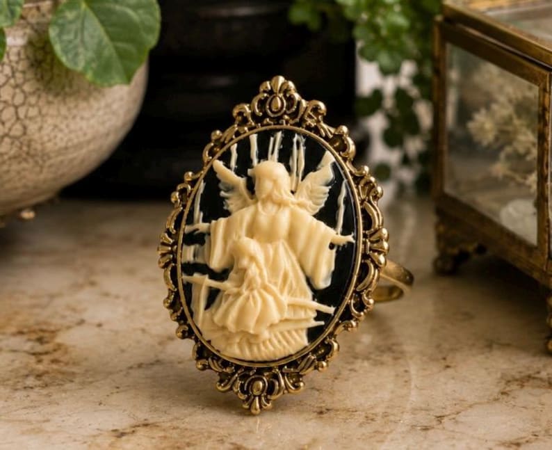 K&ouml;nnte beinhalten: Antik anmutender Ring mit ovaler Kamee, die eine cremefarbene Engelsfigur auf schwarzem Hintergrund zeigt. Der Ring hat einen goldfarbenen Filigranrahmen und -band, der eine Vintage-&Auml;sthetik zeigt. Der Ring liegt auf einer Marmoroberfl&auml;che.