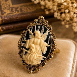 K&ouml;nnte beinhalten: Goldfarbener Ring mit ovaler Cameo-Darstellung eines cremefarbenen Engels und Kindes vor schwarzem Hintergrund. Der Ring hat eine verzierte, filigrane Fassung und einen schlichten Ring. Der Ring liegt auf einer beigen Samtoberfl&auml;che.
