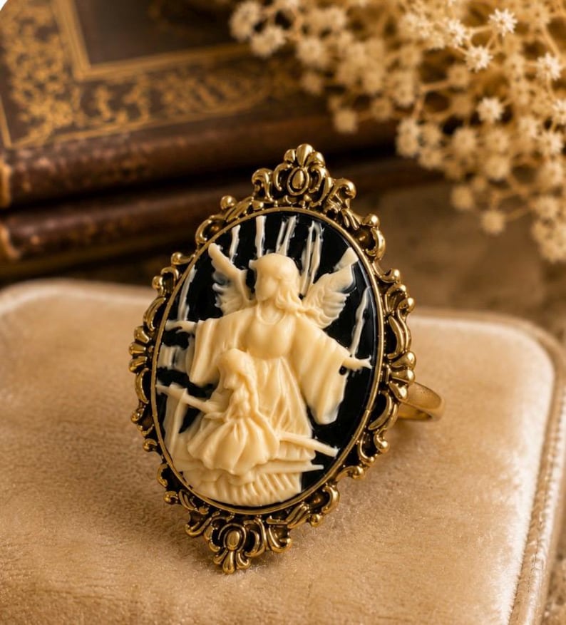 K&ouml;nnte beinhalten: Goldfarbener Ring mit ovaler Cameo-Darstellung eines cremefarbenen Engels und Kindes vor schwarzem Hintergrund. Der Ring hat eine verzierte, filigrane Fassung und einen schlichten Ring. Der Ring liegt auf einer beigen Samtoberfl&auml;che.