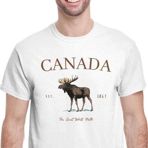 May include: White t-shirt featuring a brown moose illustration with the word "CANADA" above it. Below the moose is the text "The Great White North" and the year "1867".